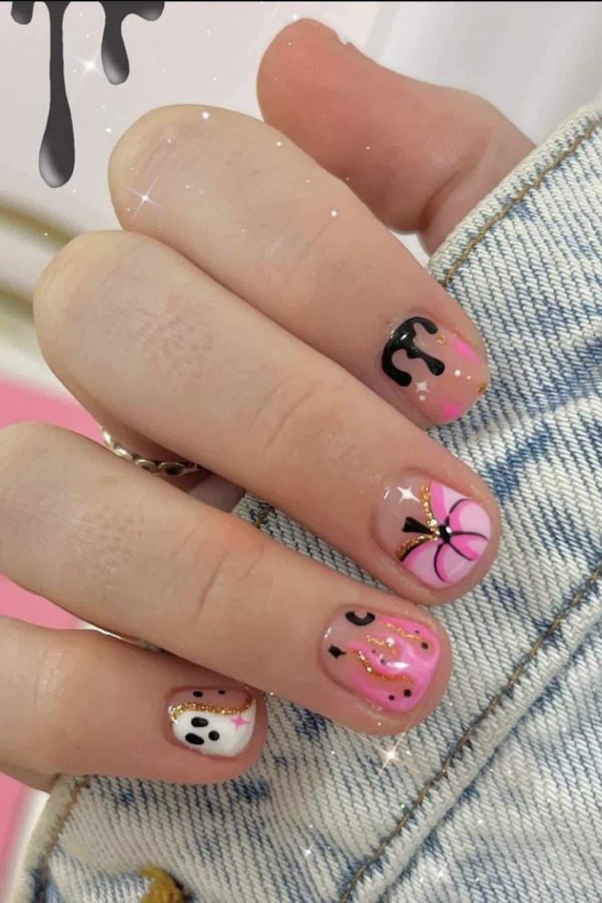 A hand with short nails painted pink, black, and white, featuring designs like a bow, drips, and dots, resting on denim fabric.