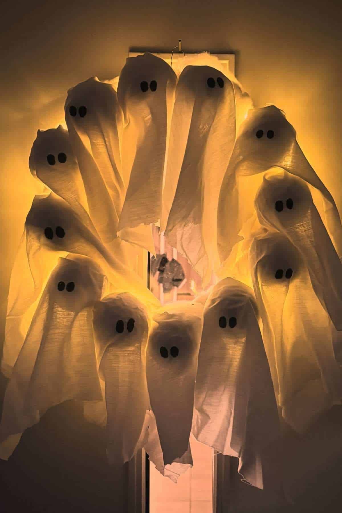 Nine ghost decorations made from white cloth and black eyes are arranged in a circle on a door, illuminated by warm yellow light.