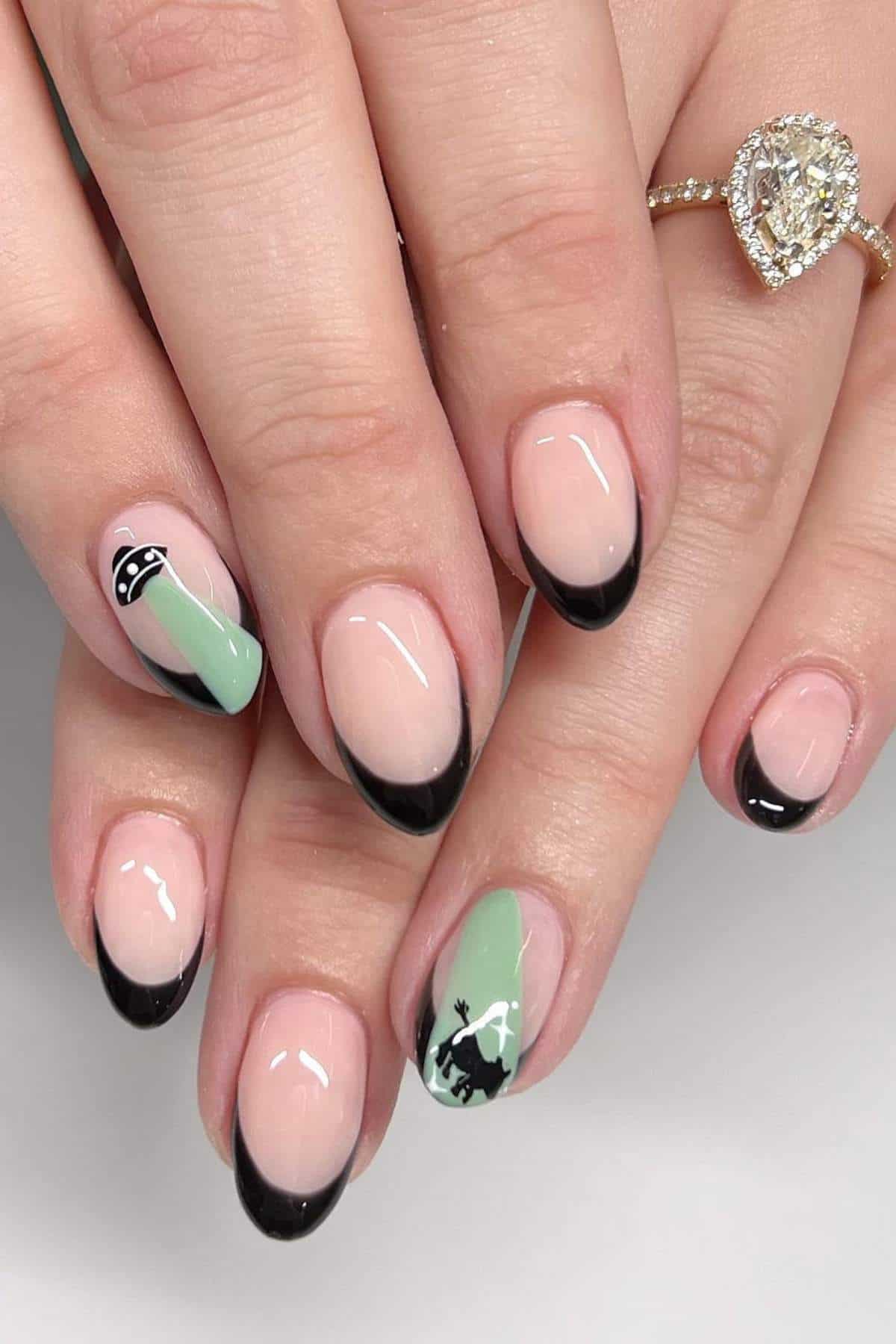 Close-up of manicured hands with black French tips; one nail features a green accent with a small black silhouette of a figure and a UFO.