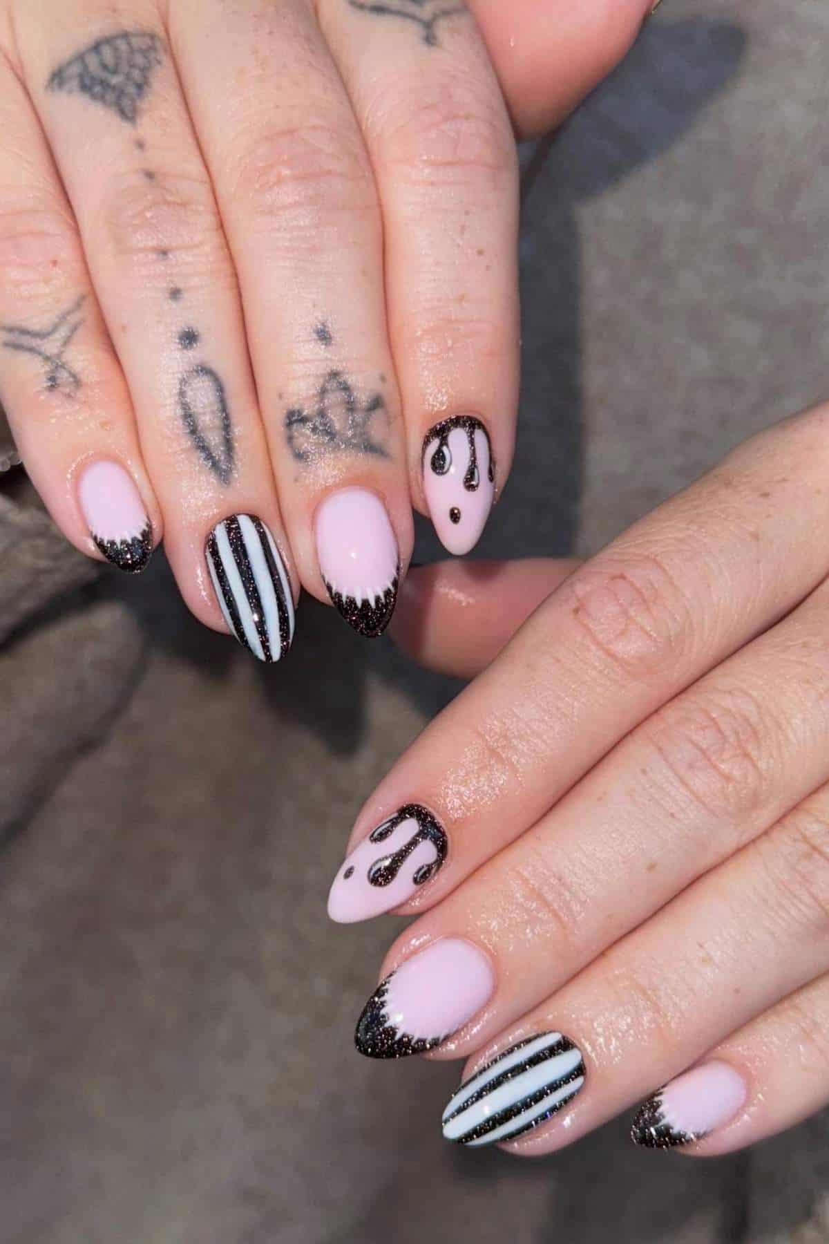 Hands with tattoos showing manicured nails featuring pink polish with black squiggles, black glitter tips, and black-and-white vertical stripes on some fingers.