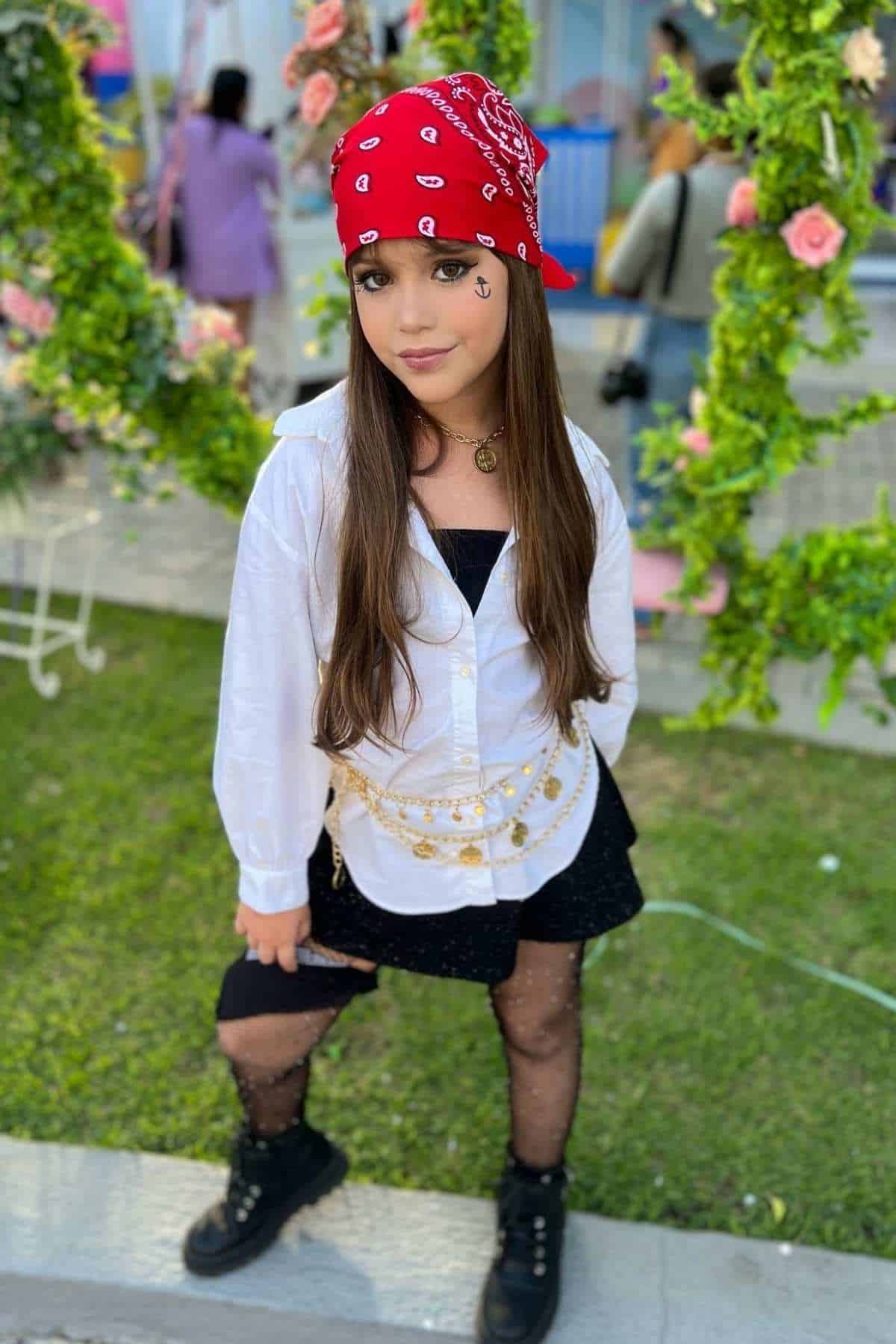 Young girl wearing a red bandana, white shirt, black skirt, patterned tights, and boots poses outdoors with greenery and flowers in the background.
