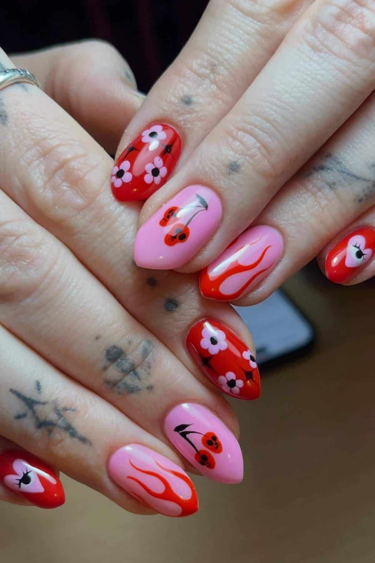 Hands with long almond-shaped nails painted in pink and red, featuring designs of cherries, flames, and small flowers.