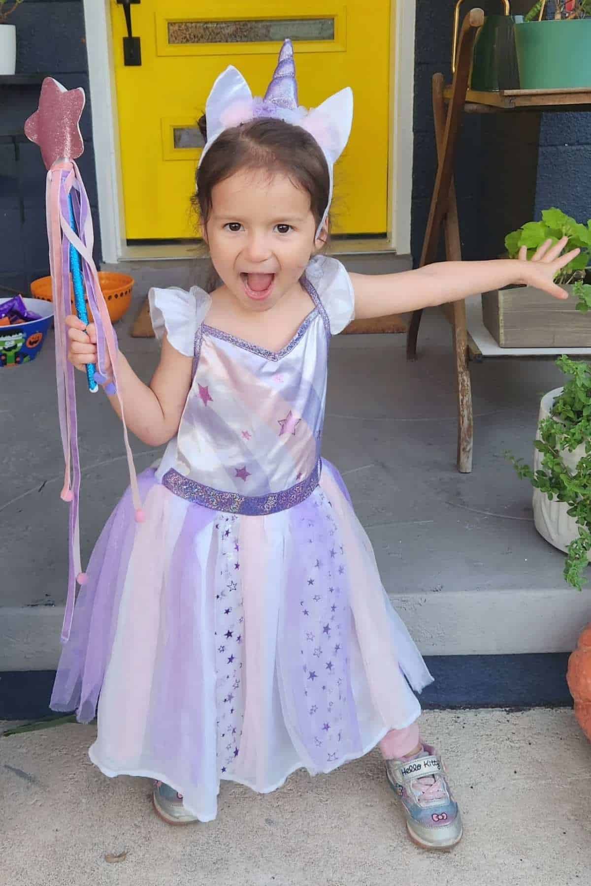 Young child in a unicorn costume stands on a porch, holding a star wand and smiling with arms outstretched.