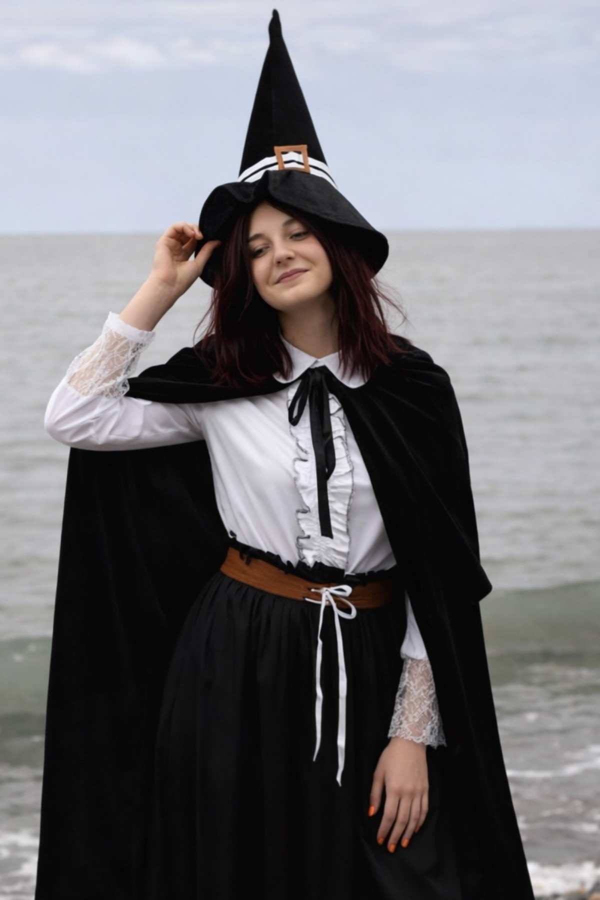 A person wearing a black witch hat, black cape, and white blouse stands by the sea, smiling with one hand touching the hat.