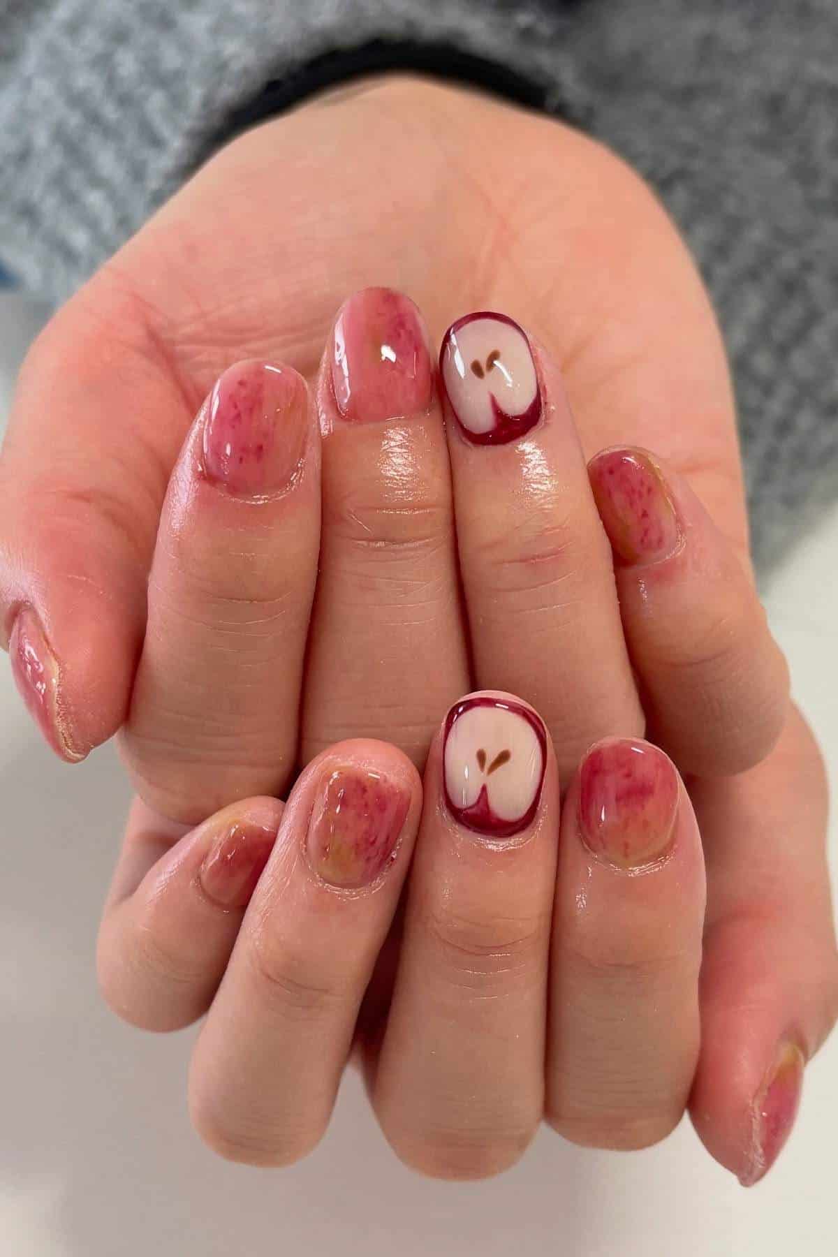 A pair of hands with short nails painted to resemble apple skin; two nails feature a design of an apple sliced in half with seeds visible.