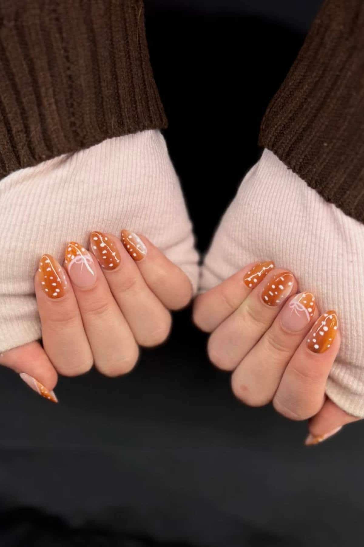 Hands with brown and white nail art featuring dots and abstract bow designs; person is wearing brown and cream long-sleeve knit tops.