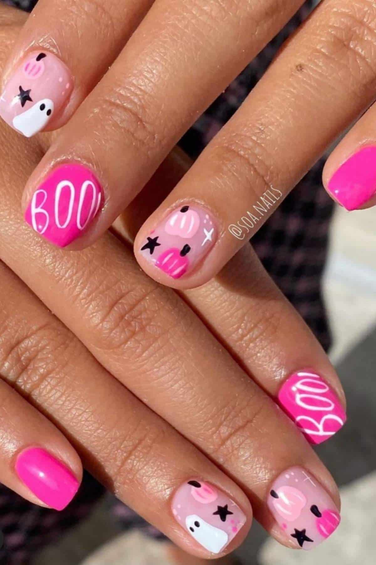 Hands with pink and nude Halloween-themed nail art, featuring the word "BOO," white ghost designs, black stars, and hearts on short, neatly manicured nails.