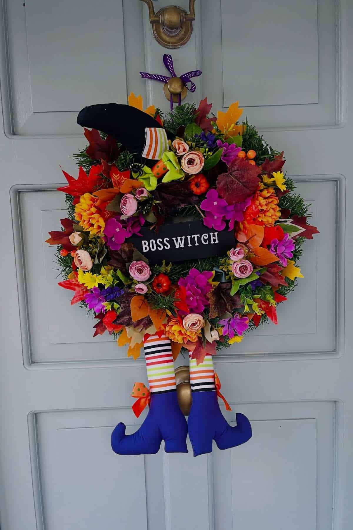 A festive wreath with fall leaves, flowers, and a "Boss Witch" sign hangs on a door, decorated with a witch hat and striped legs with purple shoes.