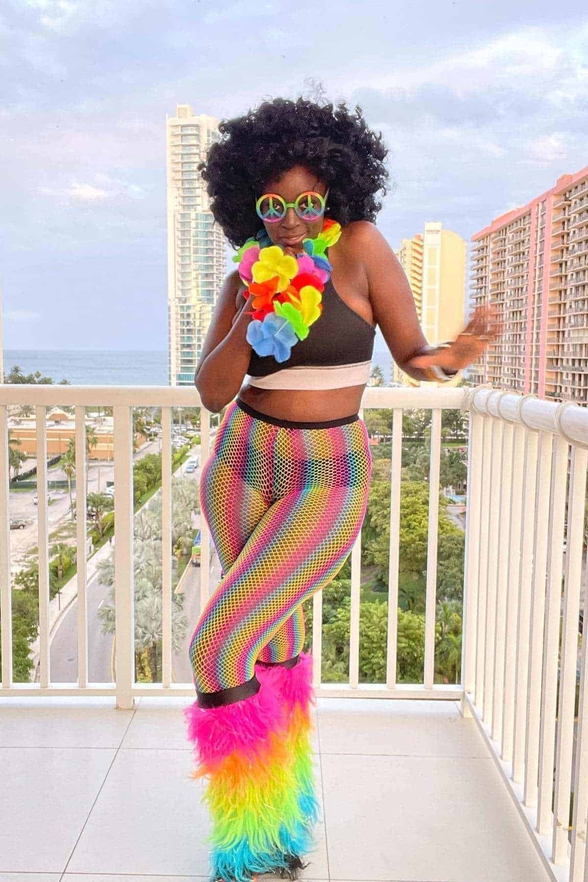 Person in rainbow bell-bottom pants, black crop top, colorful sunglasses, and furry leg warmers poses on a balcony with tall buildings and greenery in the background.