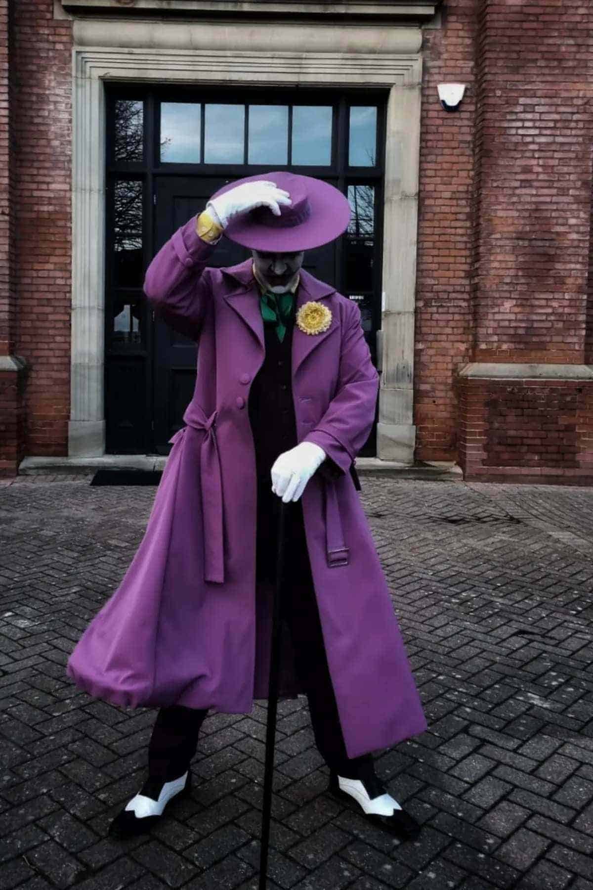 Person in a purple coat, matching hat, white gloves, and makeup poses with a cane outside a brick building, tilting their hat downward.