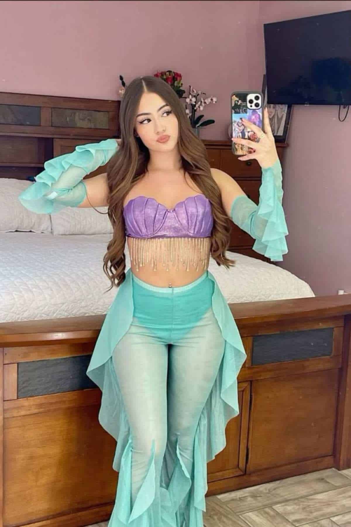 Woman in a purple top with fringe and turquoise ruffled pants takes a mirror selfie in a bedroom with wooden furniture and a wall-mounted TV.