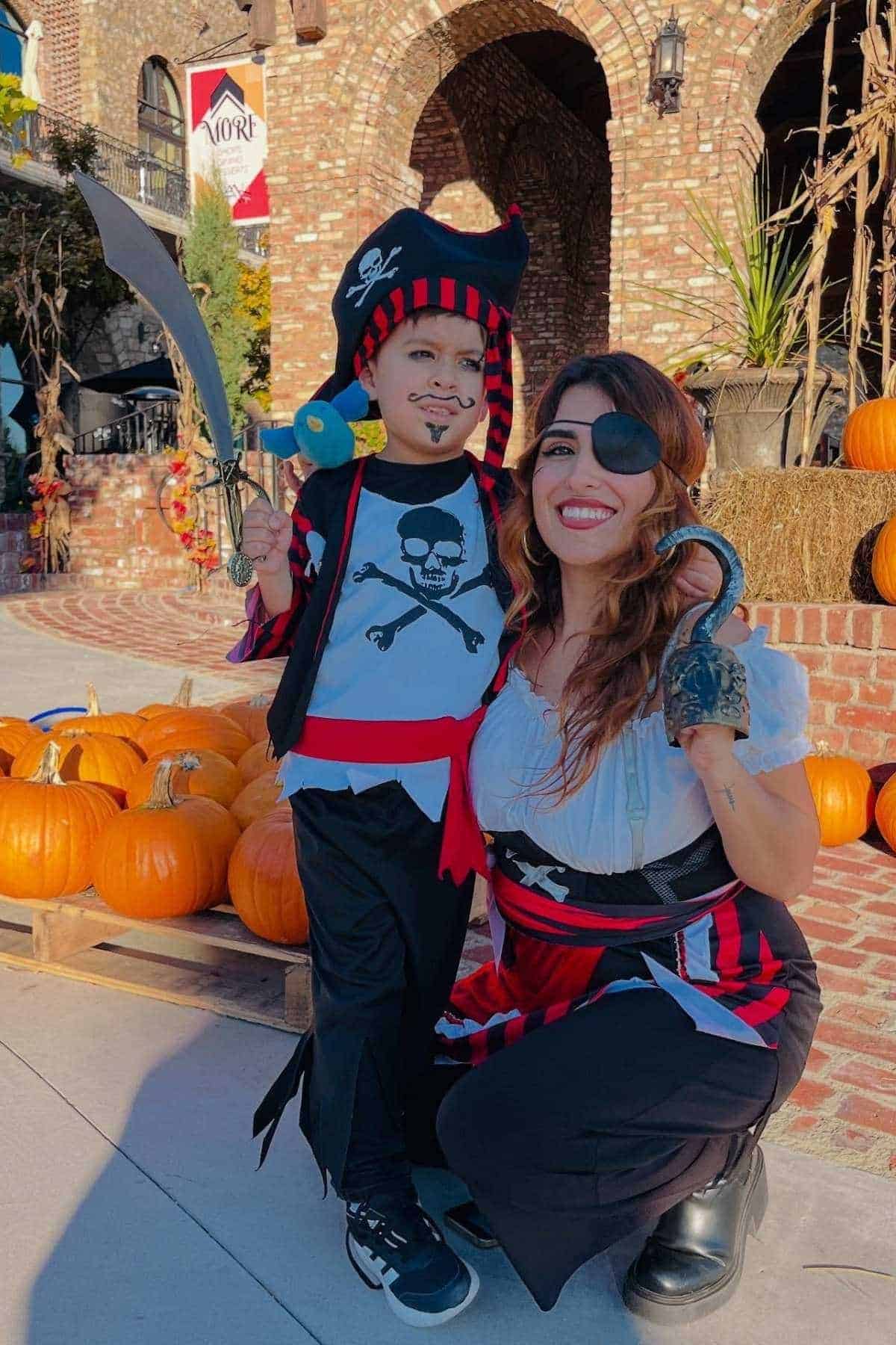 An adult and child dressed as pirates pose and smile in front of a display of pumpkins and a brick building decorated for autumn.