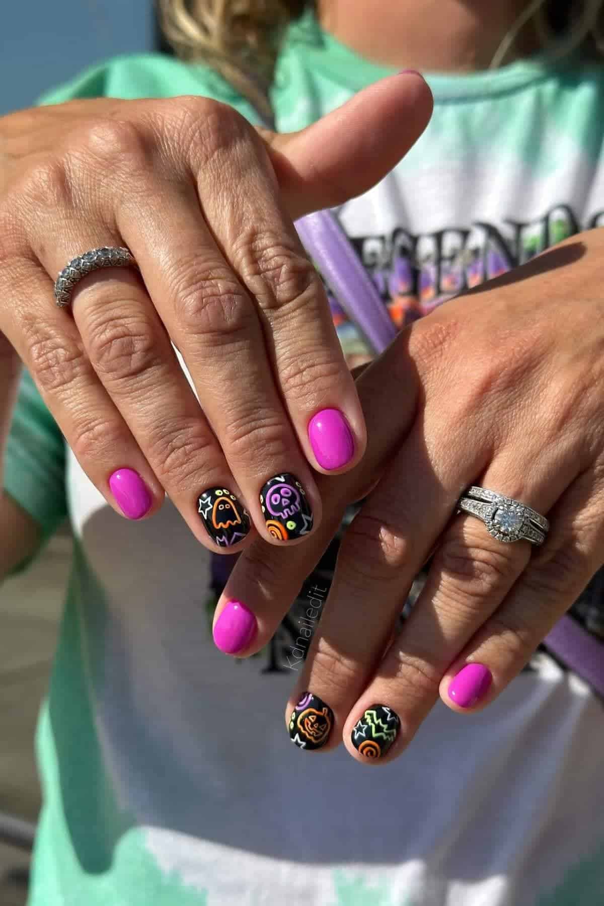 Hands with bright pink and Halloween-themed nail art featuring ghosts and pumpkins. Person is wearing two rings on each hand and a green and white shirt.