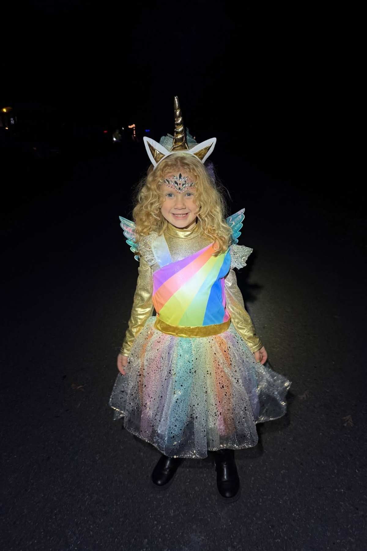 A young child in a rainbow unicorn costume with a gold horn, wings, and a sparkly skirt stands outside at night, smiling at the camera.