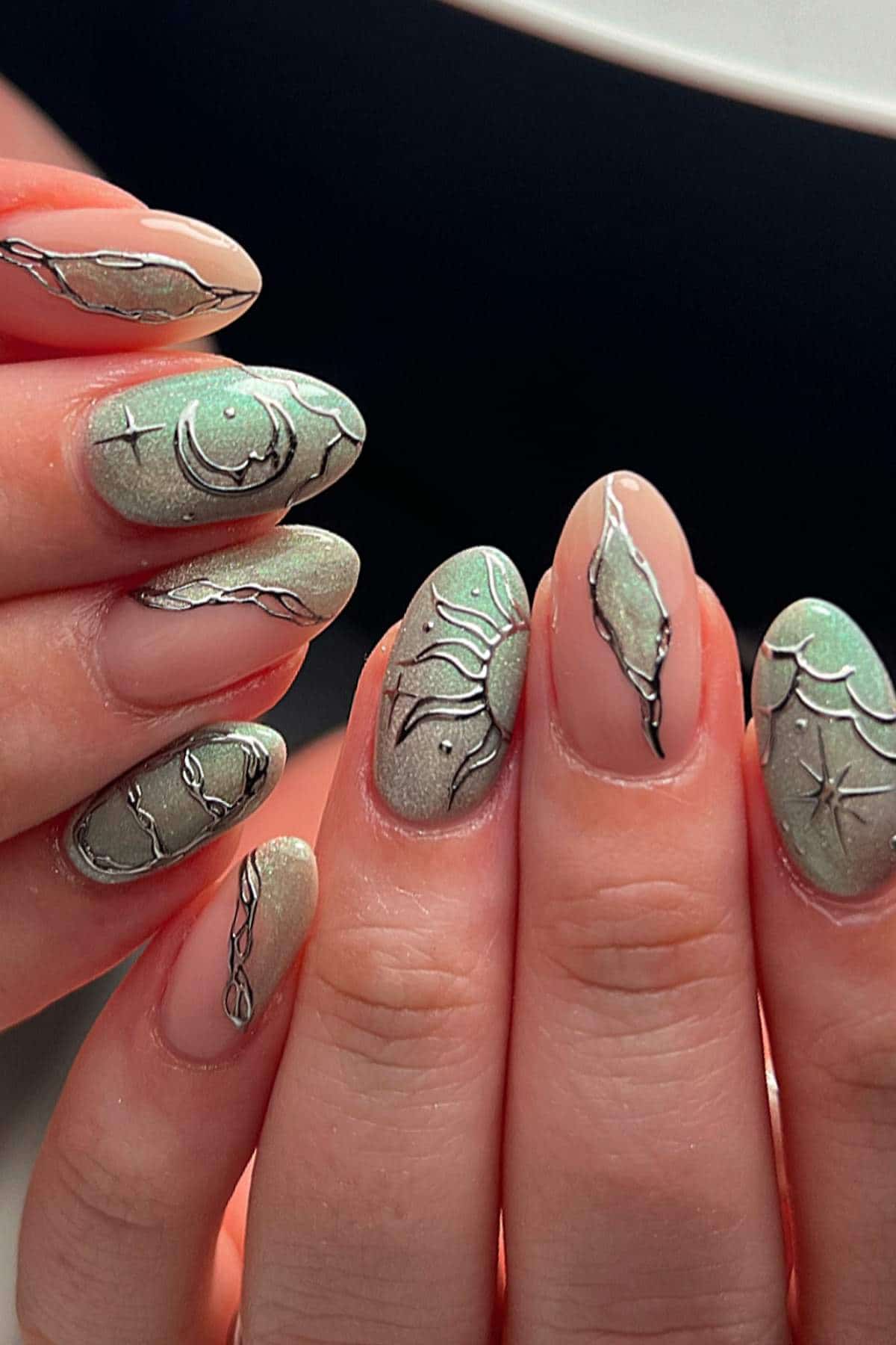 Close-up of hands with almond-shaped nails featuring green and silver abstract designs on a nude base.