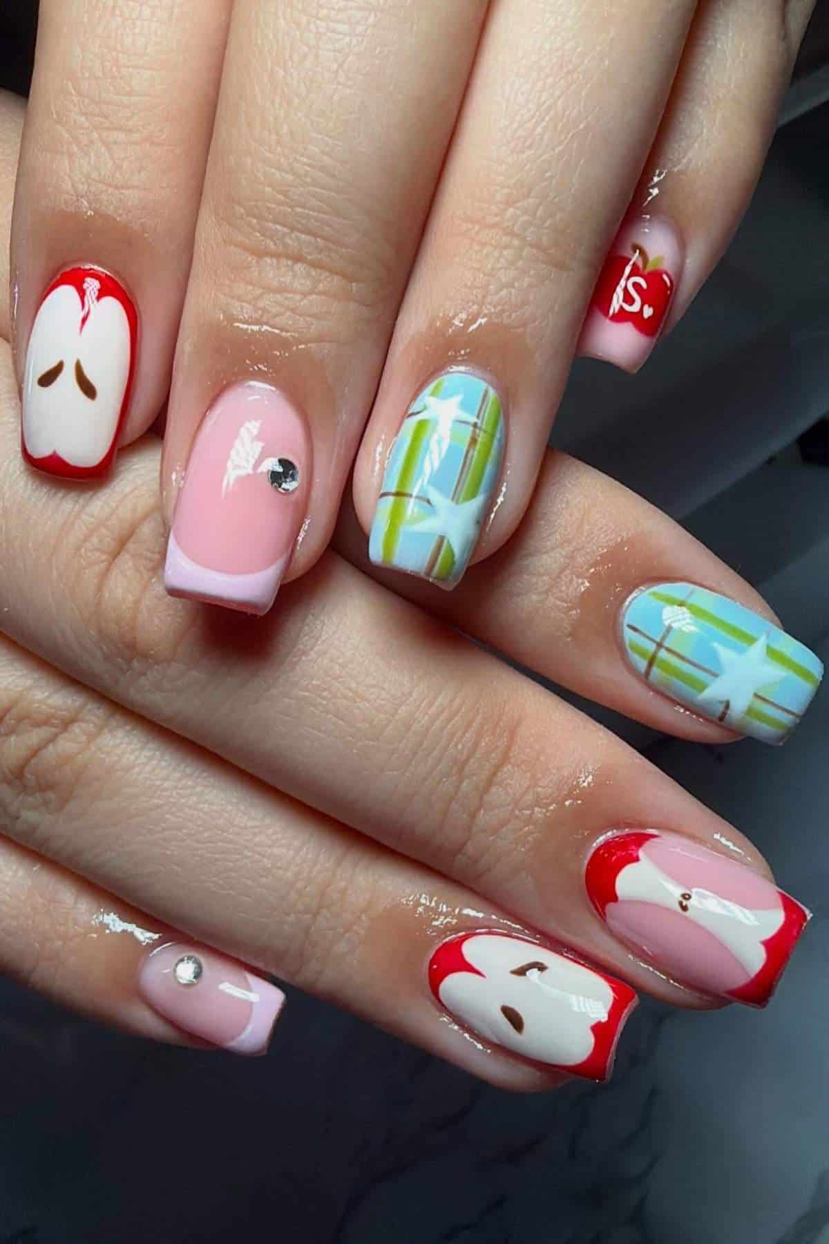 Close-up of hands with nail art featuring apple slices, plaid patterns in blue and green, and rhinestone accents on a few nails.