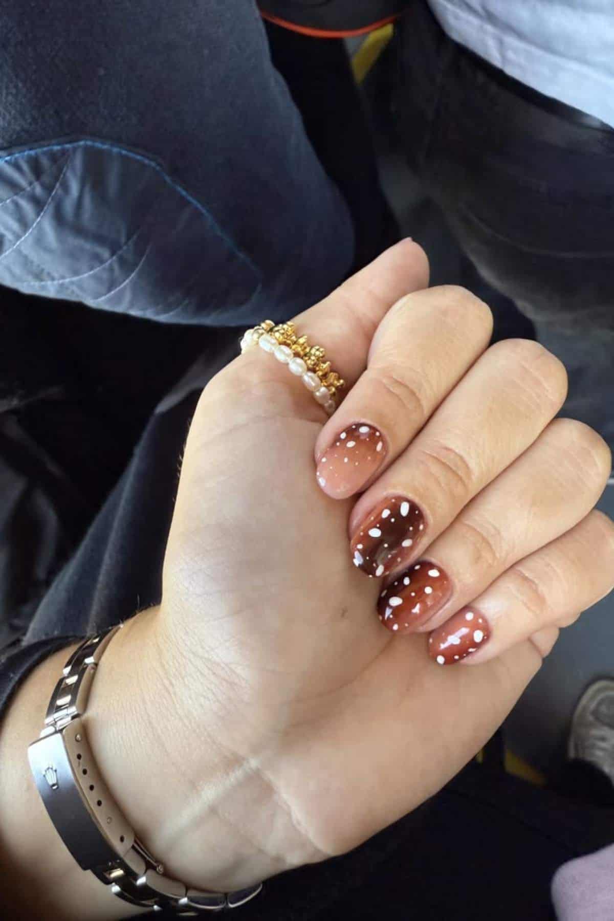 A hand with a bracelet and ring displays brown gradient nails with white polka dots. Other people’s clothing is visible in the background.