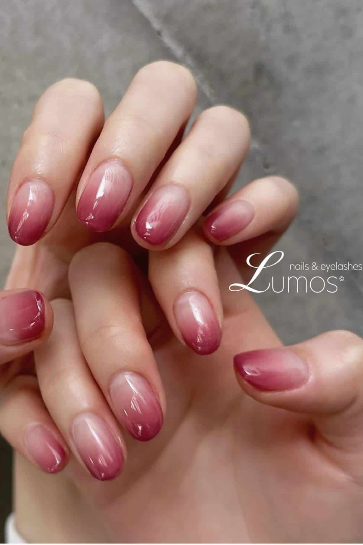 Close-up of hands with long, almond-shaped nails featuring a pink ombre manicure. The background is blurred and features the “Lumos nails & eyelashes” logo.