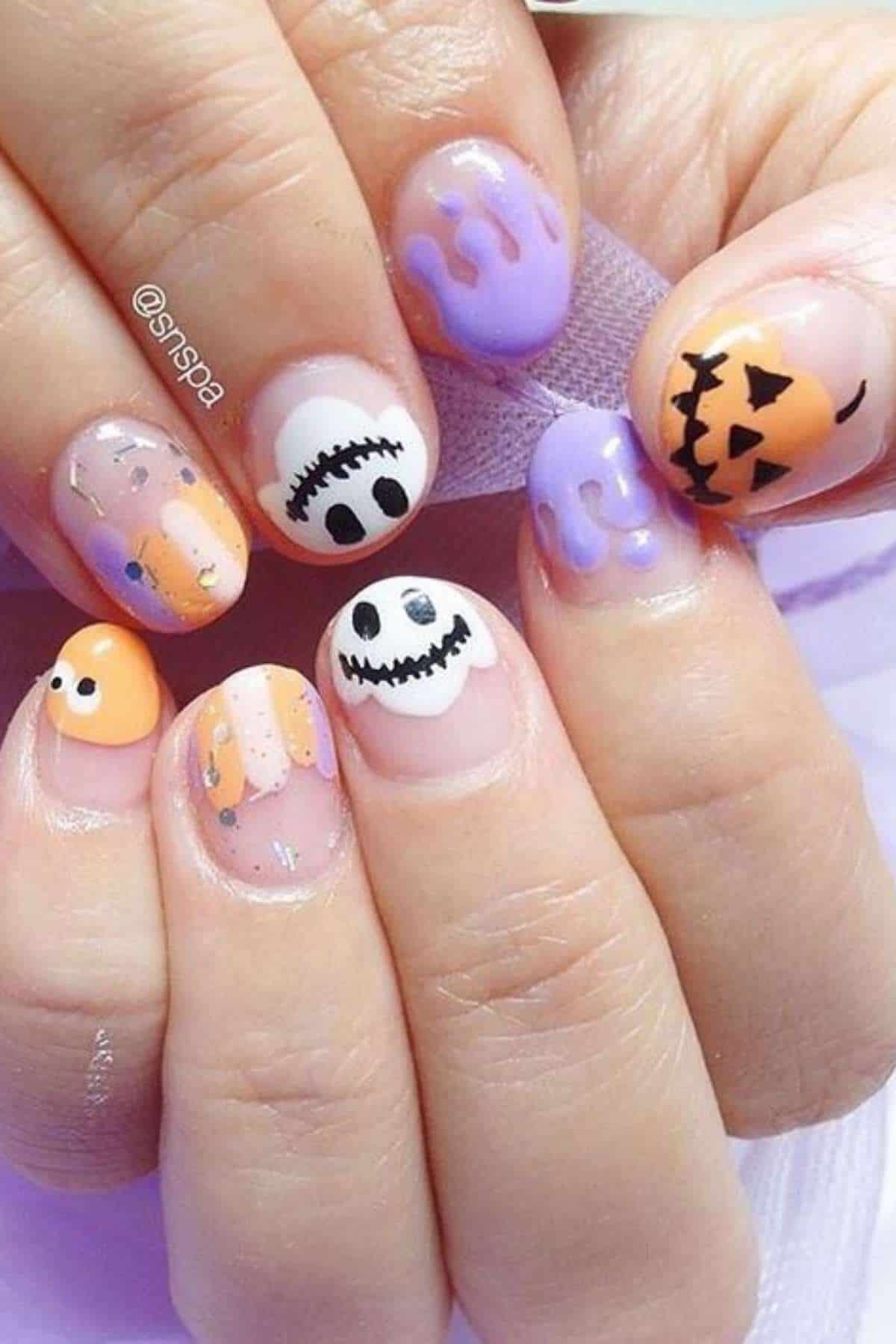 Fingernails with Halloween-themed nail art, including a pumpkin face, ghost, stitched mouth, and purple dripping designs on a mix of orange and lavender backgrounds.