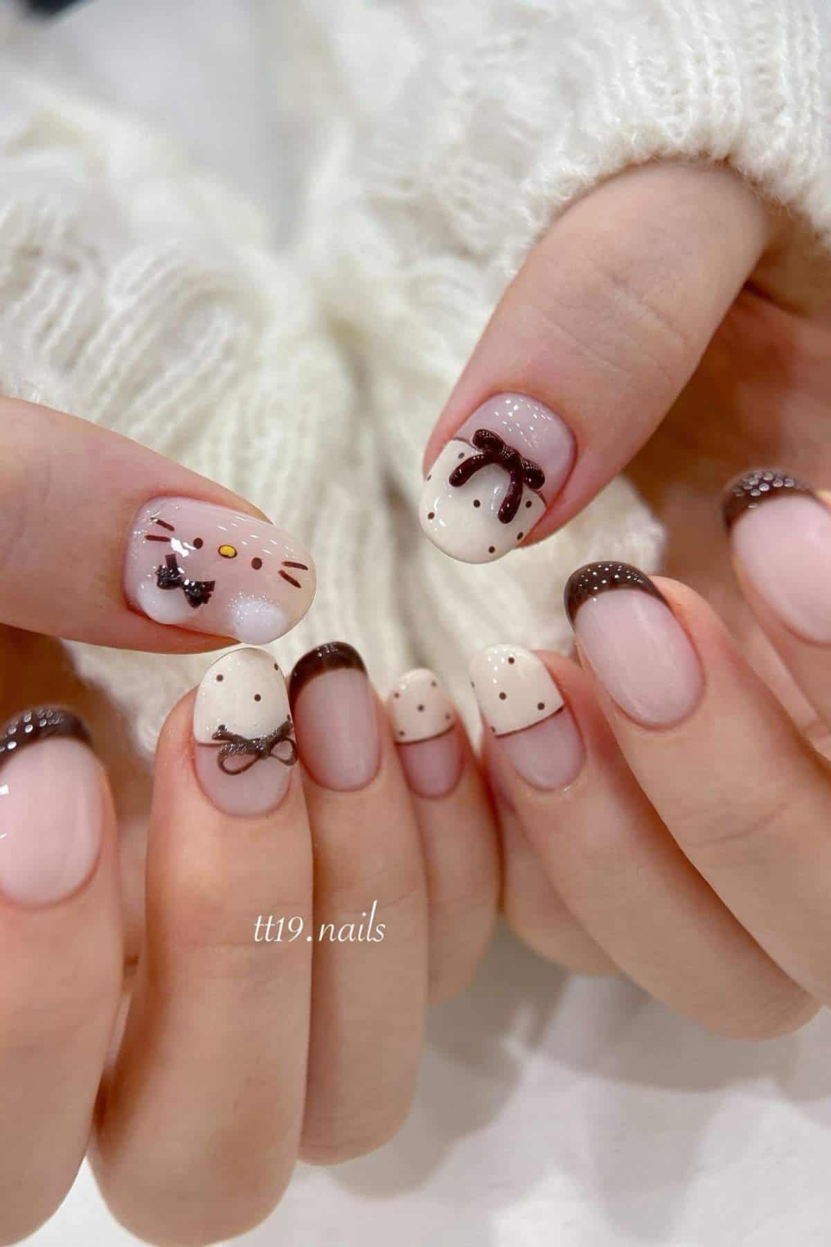 Hands with short, almond-shaped nails featuring brown French tips, polka dots, and cartoon cat faces with bows, against a white knit sweater background.