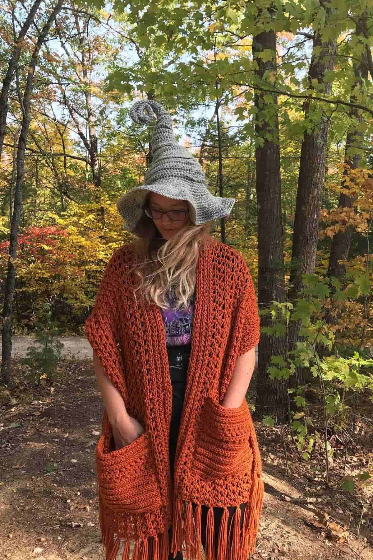 A person wearing a gray pointed hat and an orange knit shawl stands in a forest with autumn foliage.
