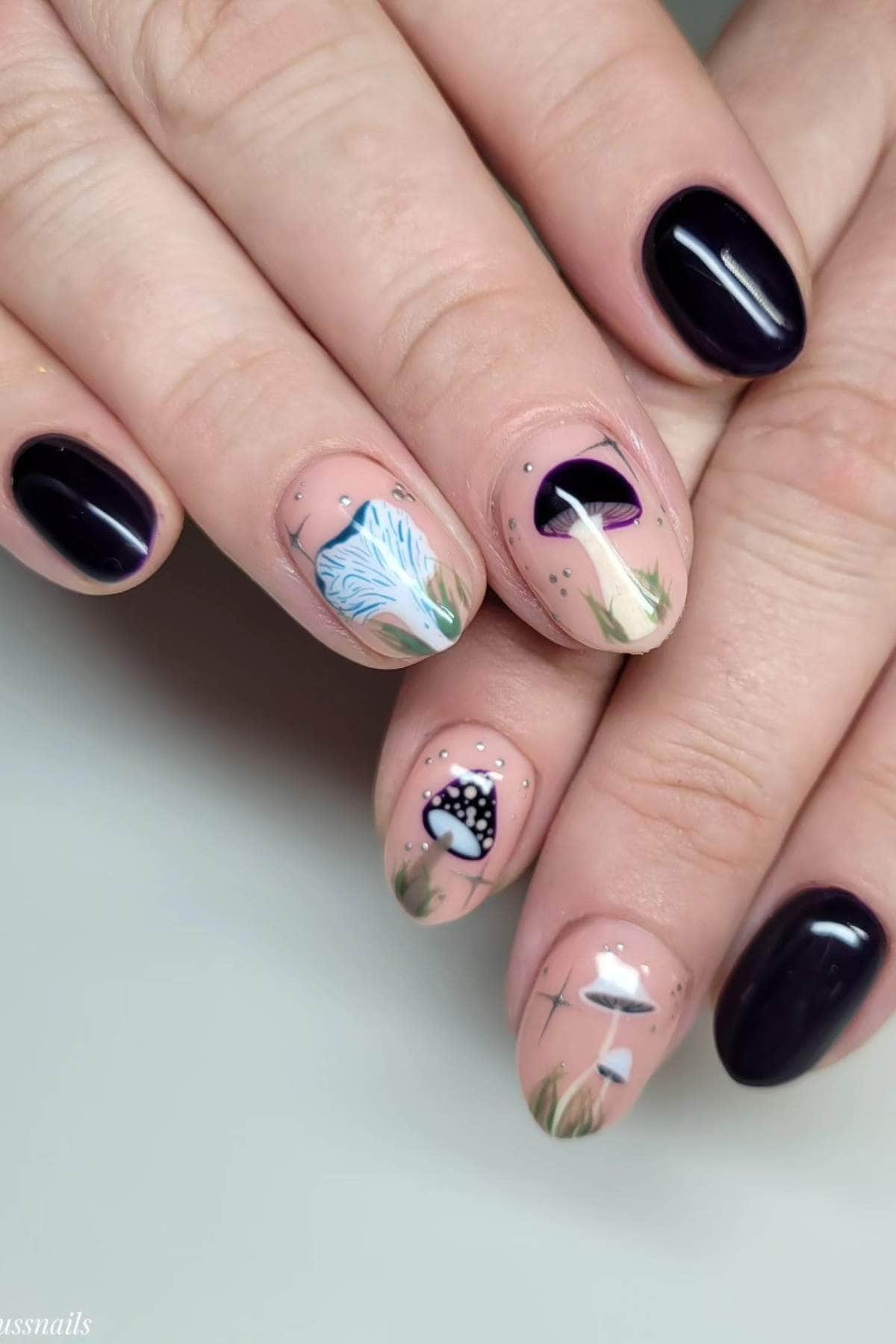 A hand with black and nude nail polish; some nails feature detailed designs of mushrooms, grass, and abstract art.