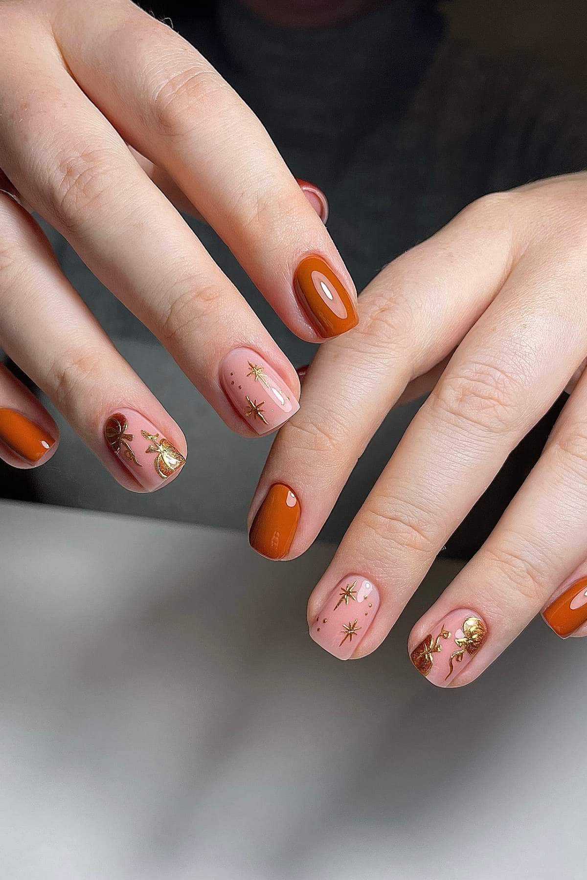 Hands with manicured nails; some nails are painted solid burnt orange while others are nude with gold star and swirl designs.