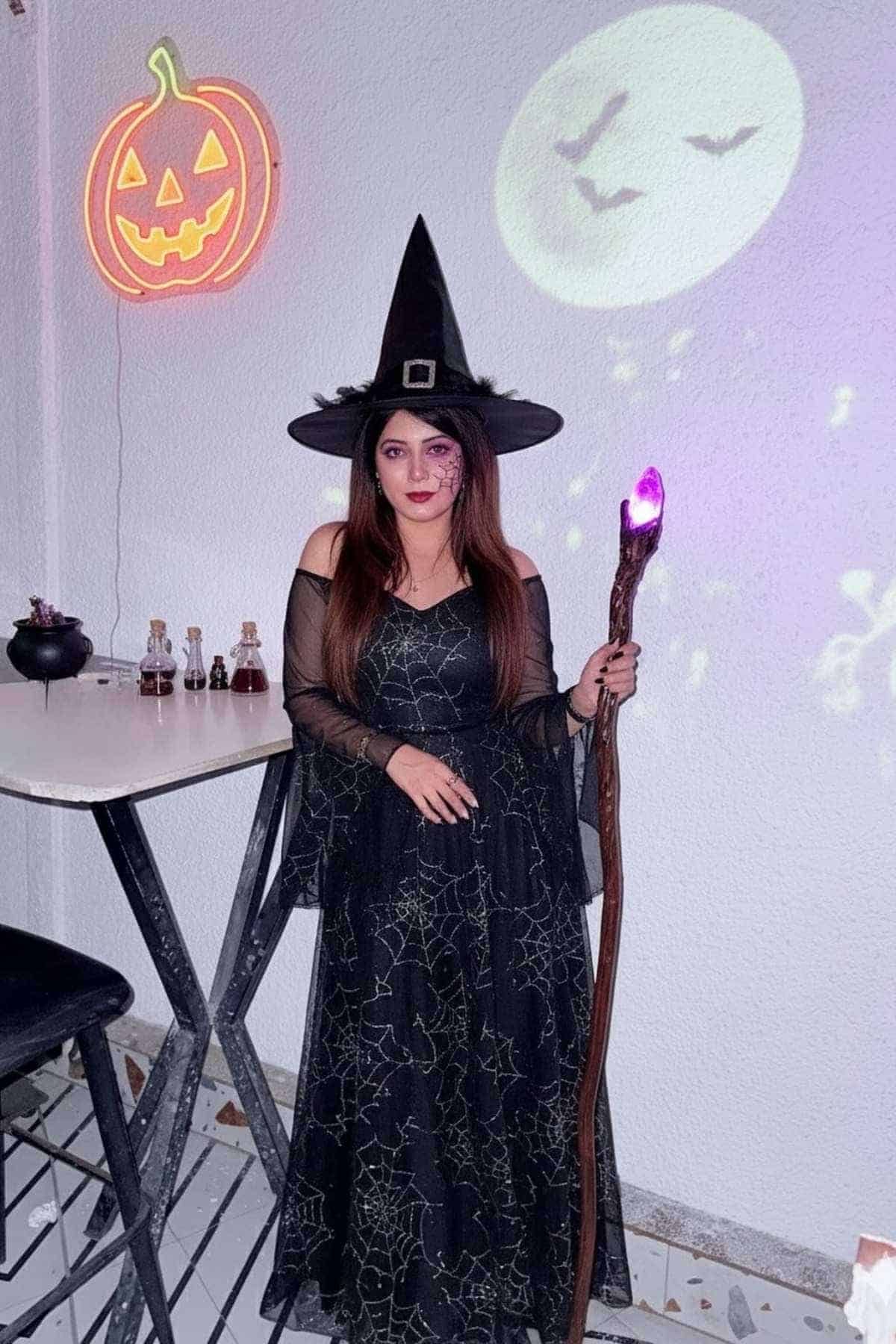 Woman in a black witch costume with a pointed hat and glowing staff stands by a table with potion bottles, with Halloween decorations in the background.