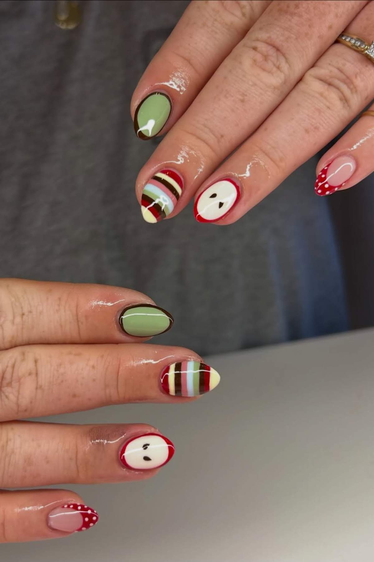 A hand with almond-shaped nails featuring designs of apples, green polish, red and white stripes, and red tips with white polka dots on a light background.