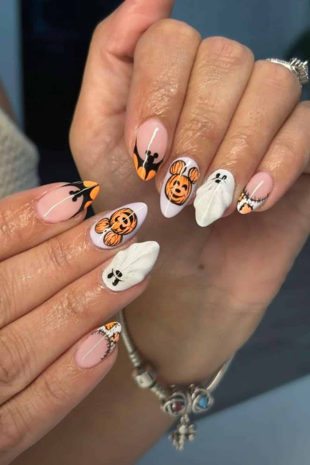 Hands with Halloween-themed nail art featuring pumpkins, ghosts, and stitched designs in orange, black, and white on almond-shaped nails.