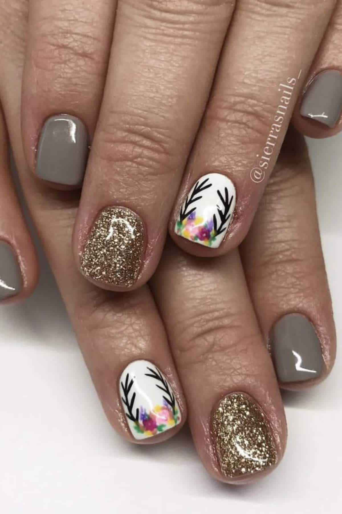 Hands with manicured nails featuring gray polish, gold glitter, and white accent nails with colorful floral and black antler designs.