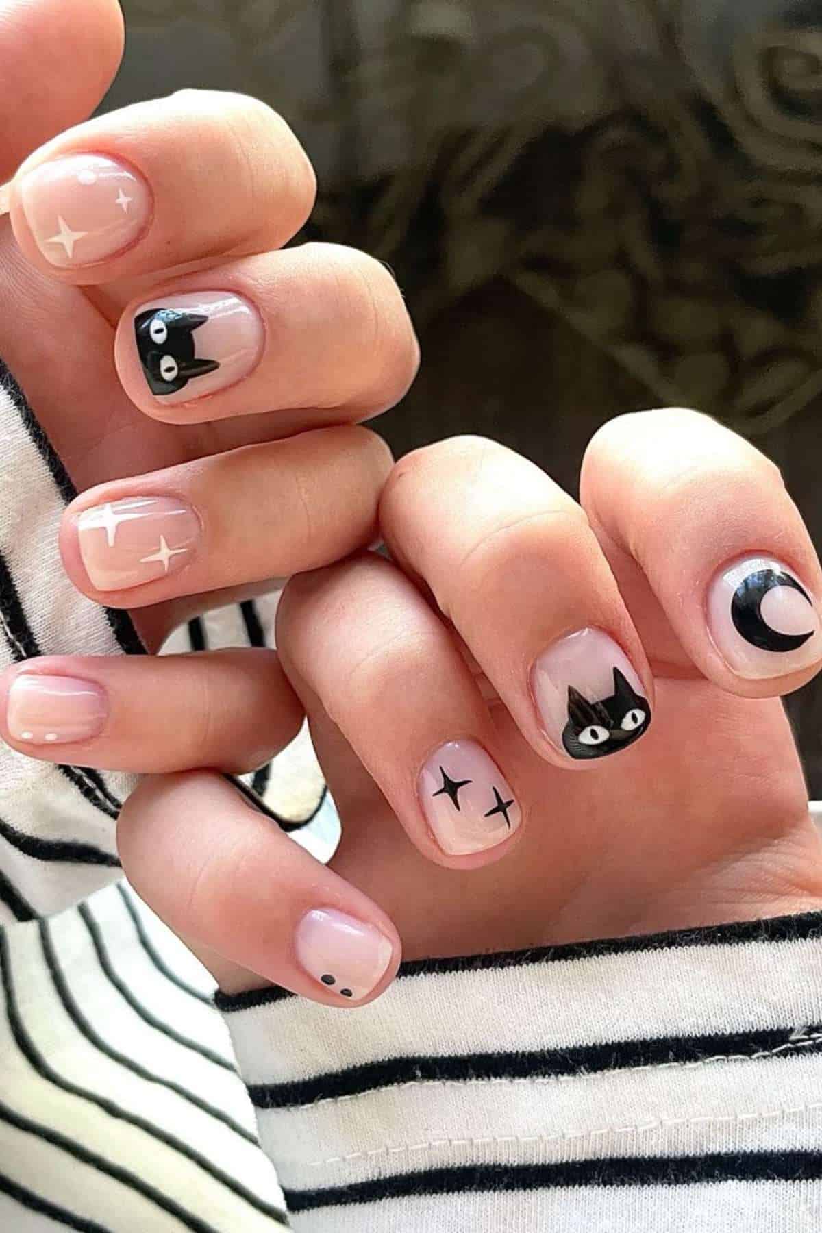 Hands with short, nude nails featuring black cat faces, stars, a crescent moon, and dots as nail art. The person wears a black and white striped long-sleeve shirt.