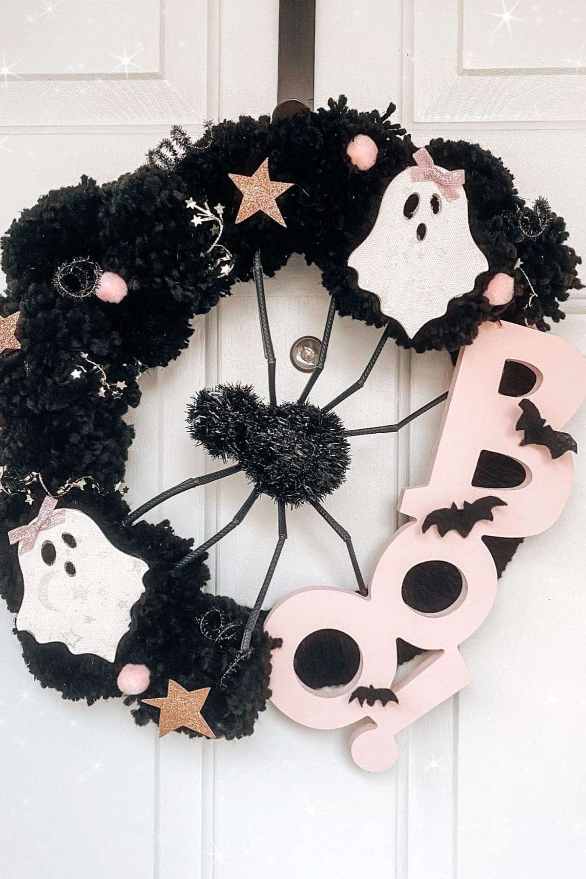 A black Halloween wreath with faux ghosts, pink stars, a large spider, and the word "BOO" in pink letters with black bat cutouts, hanging on a white door.