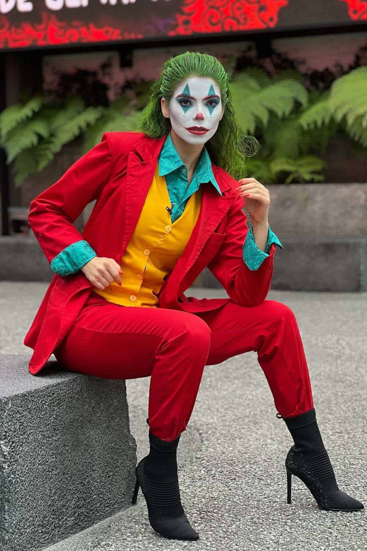 Person dressed as the Joker, wearing a red suit, yellow vest, green shirt, and black high-heeled boots, with green hair and white face paint, sitting on a concrete bench outdoors.