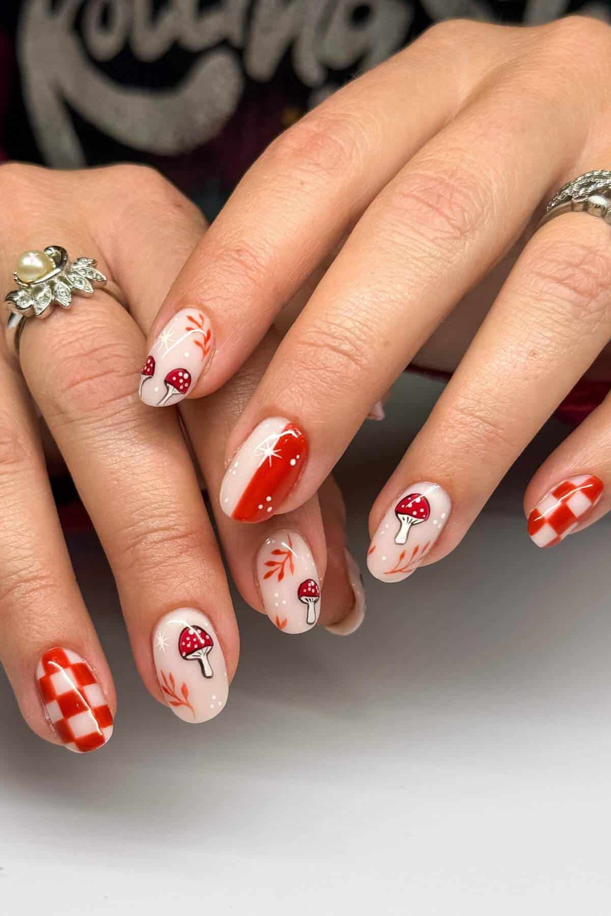 Hands with almond-shaped nails featuring red and white designs, including mushrooms, leaves, and checkerboard patterns. Person wears rings on index and ring fingers.