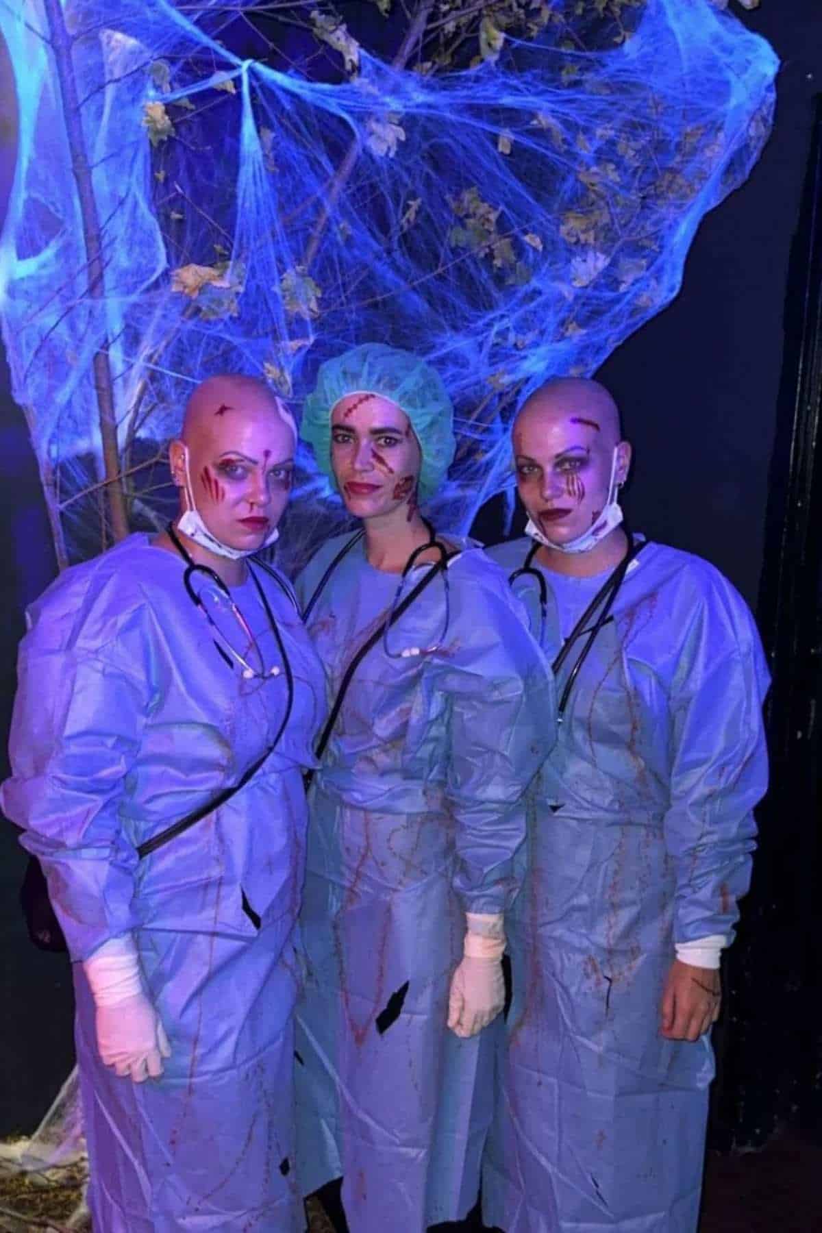 Three people in blue medical scrubs and bald caps with fake blood and wounds pose under fake spiderweb decorations.