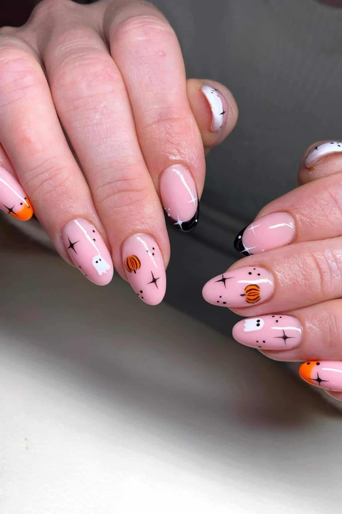 A close-up of hands with almond-shaped nails featuring Halloween-themed nail art, including ghosts, spider webs, pumpkins, dots, and black tips on a light pink base.
