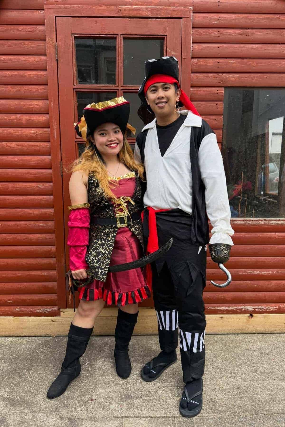 Two people in pirate costumes stand in front of a red wooden building. The woman wears a red and gold dress, while the man wears black and white clothes and holds a hook.
