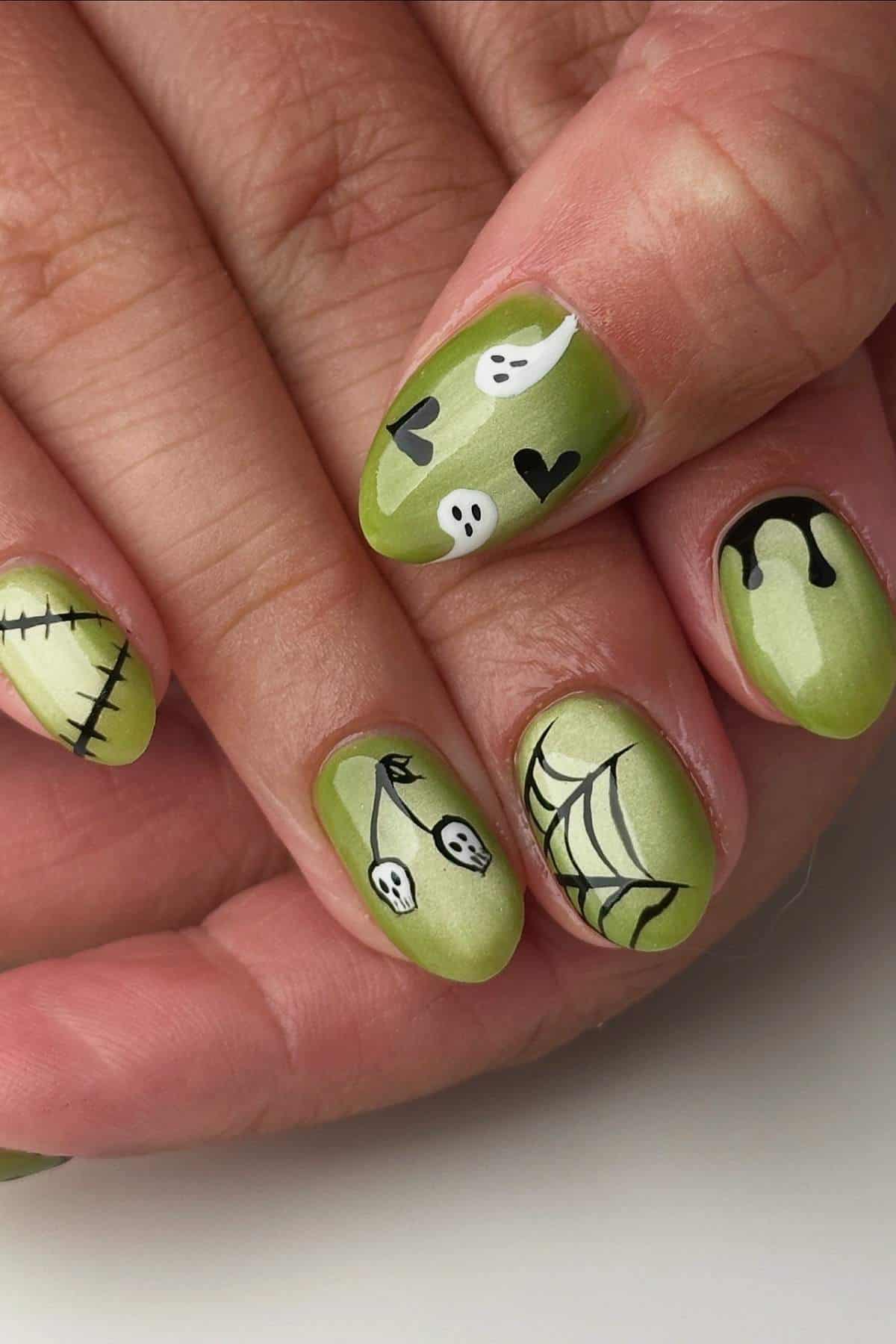 A hand with green Halloween-themed nail art featuring ghosts, hearts, stitches, drips, and a spiderweb design.