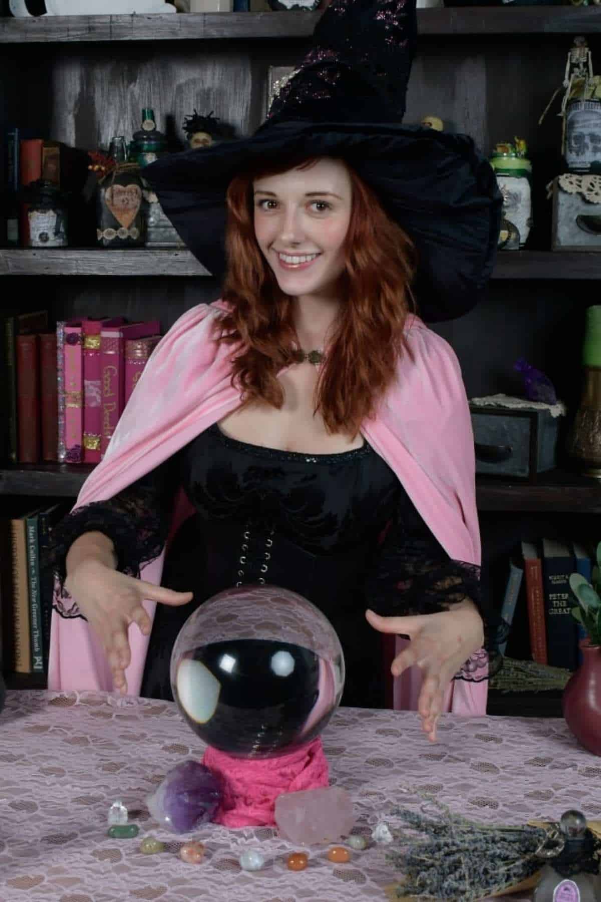 A person in a witch costume with a pink cape and hat stands behind a table with a crystal ball, stones, and herbs, in front of a bookshelf.