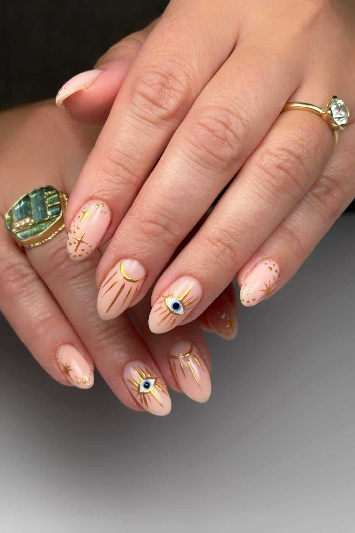 Hands with almond-shaped nails painted nude pink, featuring gold lines and blue evil eye designs; person wears gold and green rings.