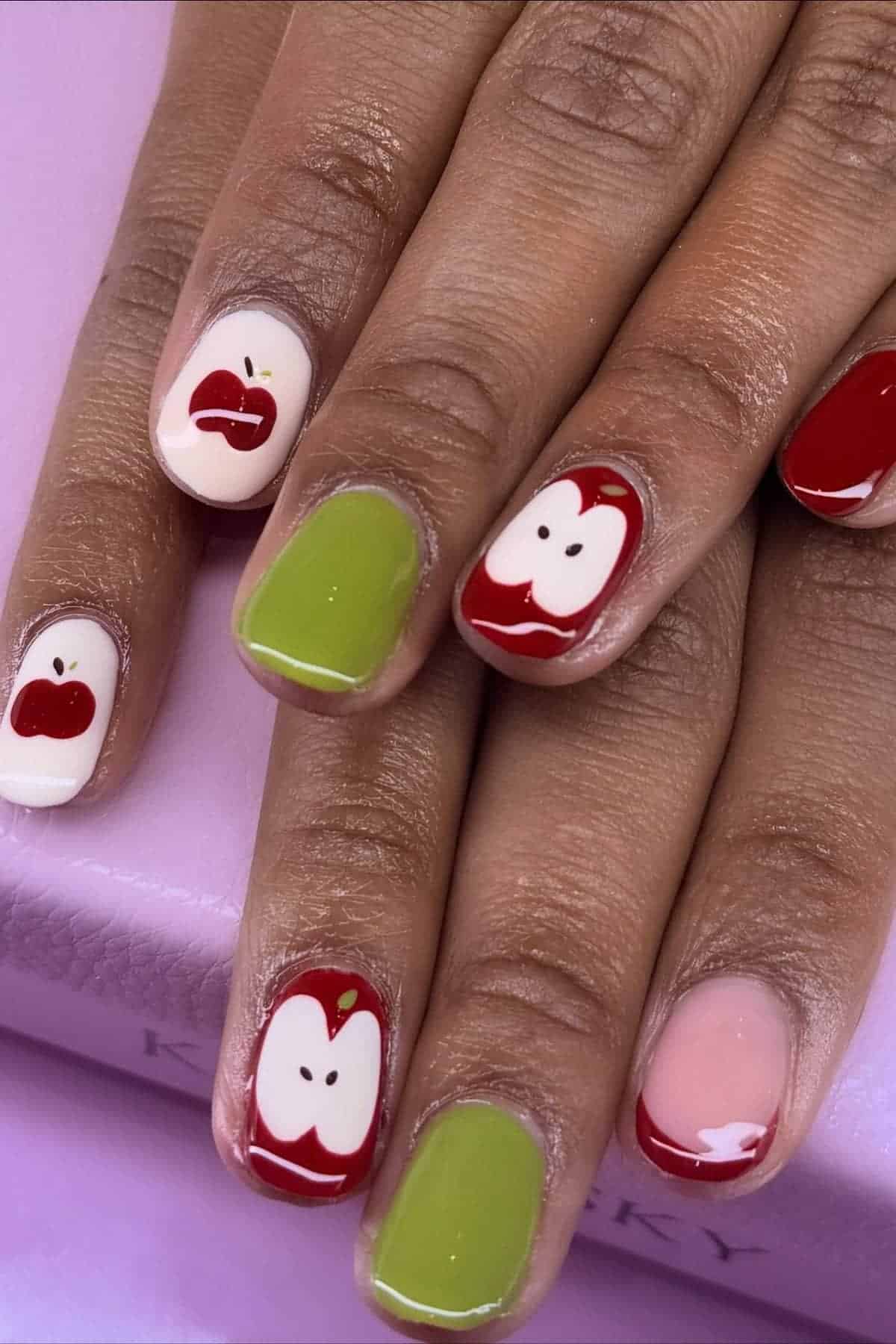 A hand with manicured nails featuring apple designs, green polish, and apple-themed nail art on a light purple surface.