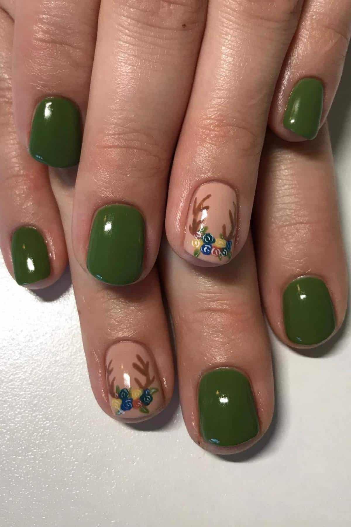Hands with short, square nails painted green; two accent nails feature nude polish with antler designs and small painted blue and yellow flowers.