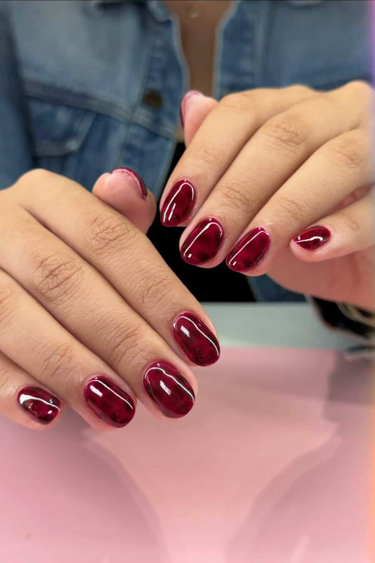 Close-up of hands with short, oval-shaped nails painted in glossy dark red polish. Person is wearing a blue denim jacket. Hands rest on a pink surface.