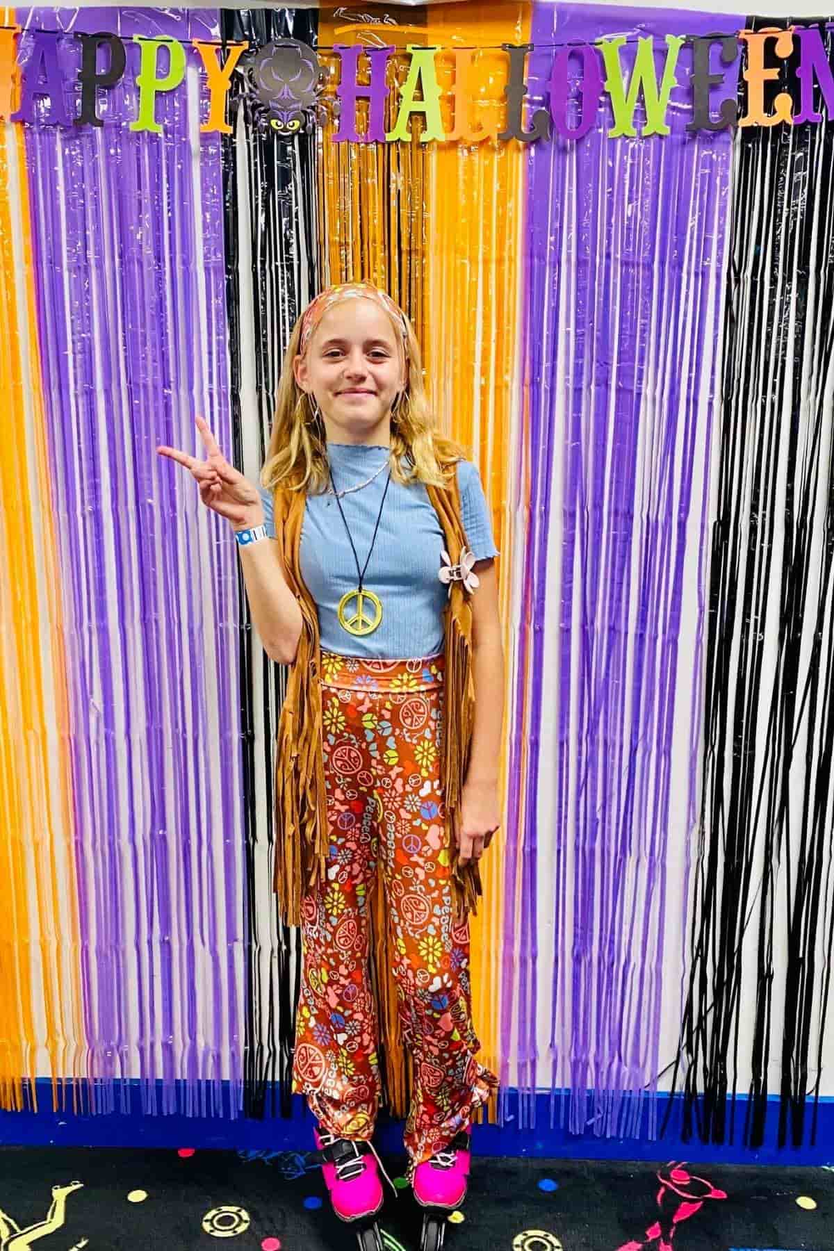 A girl in a 1970s hippie costume poses with a peace sign in front of a “Happy Halloween” banner and colorful streamers.