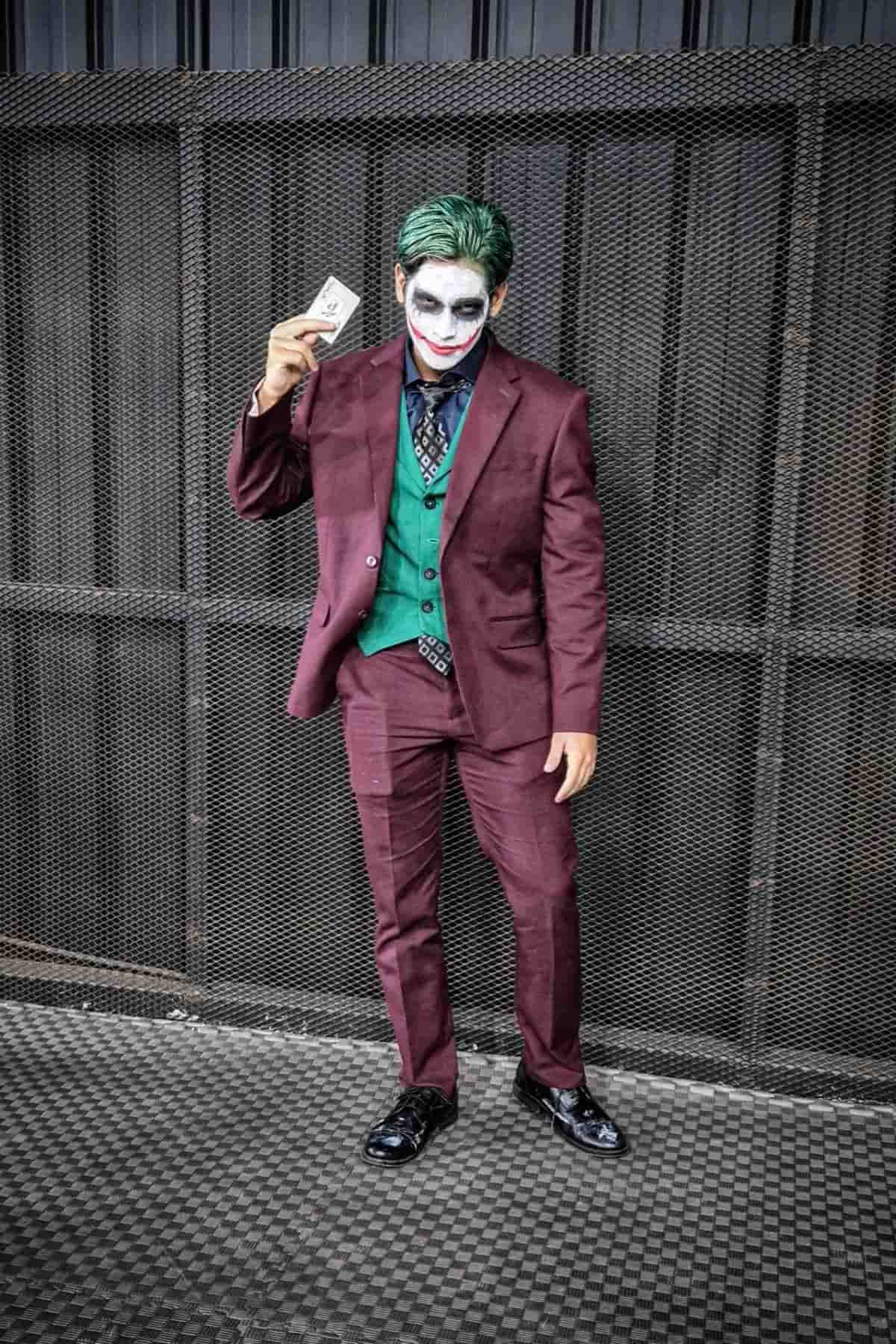Person in a burgundy suit and green vest with white face makeup and green hair holds up a playing card, standing in front of a metal fence.