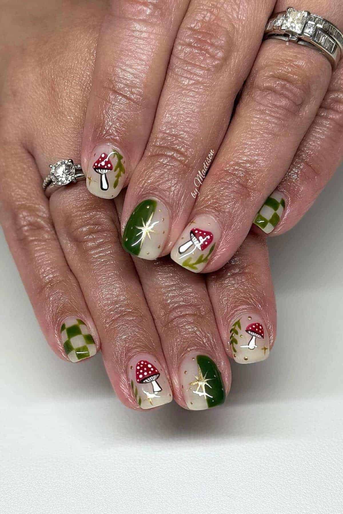 Hands with manicured nails featuring green patterns, red and white mushroom designs, starbursts, and checkered accents. Rings are worn on the ring finger.