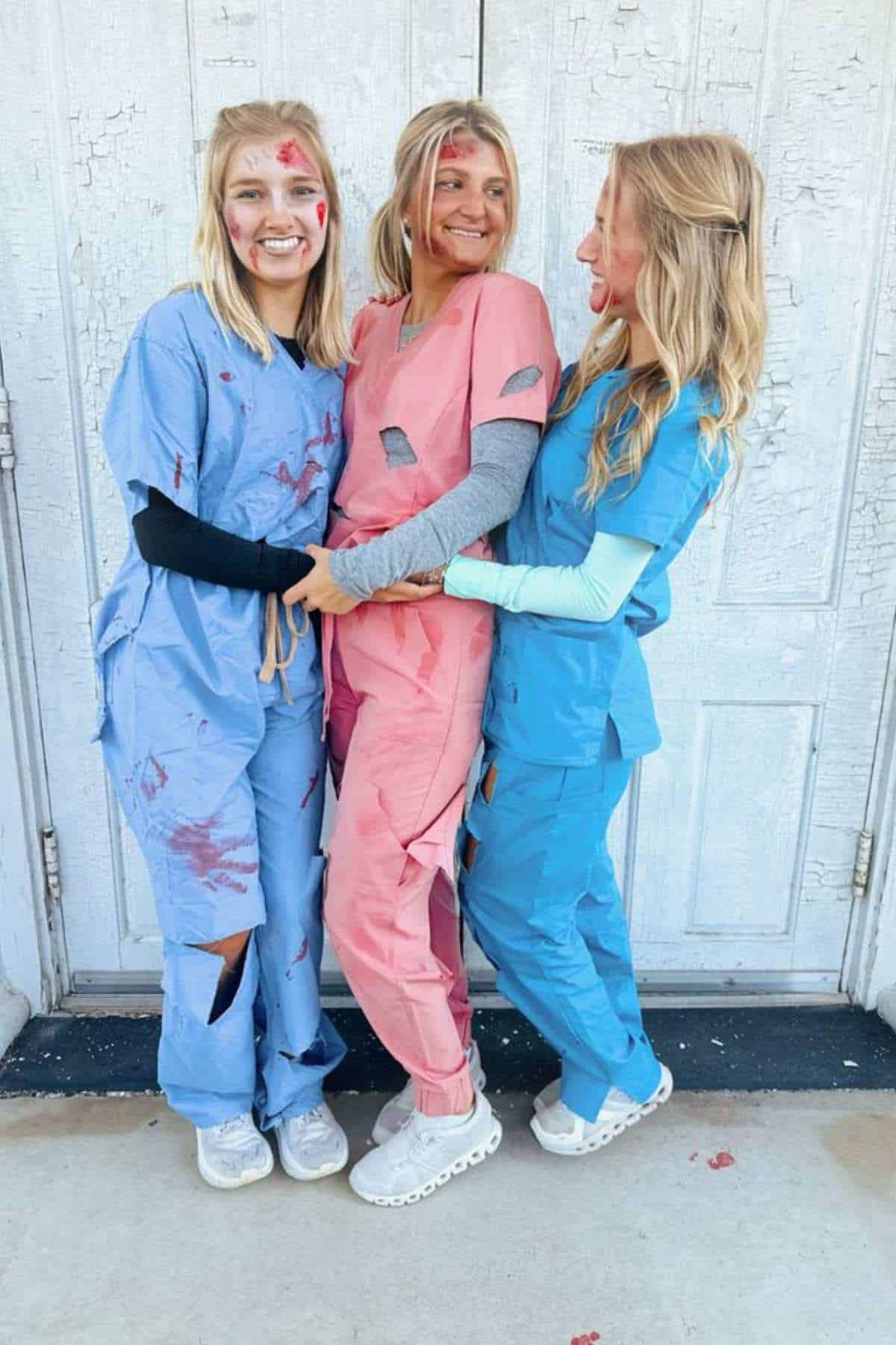 Three people in tattered scrubs with fake wounds and blood stand smiling together in front of a weathered white door.