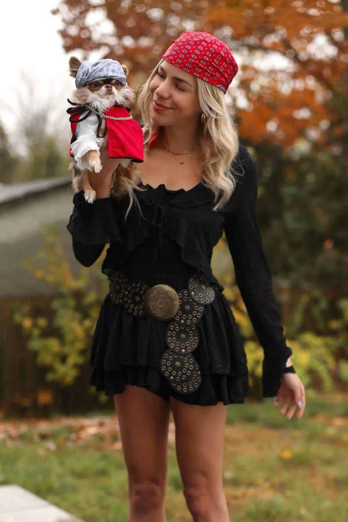 A woman in a black dress and red bandana holds a small dog dressed in a matching pirate costume outdoors with autumn trees in the background.