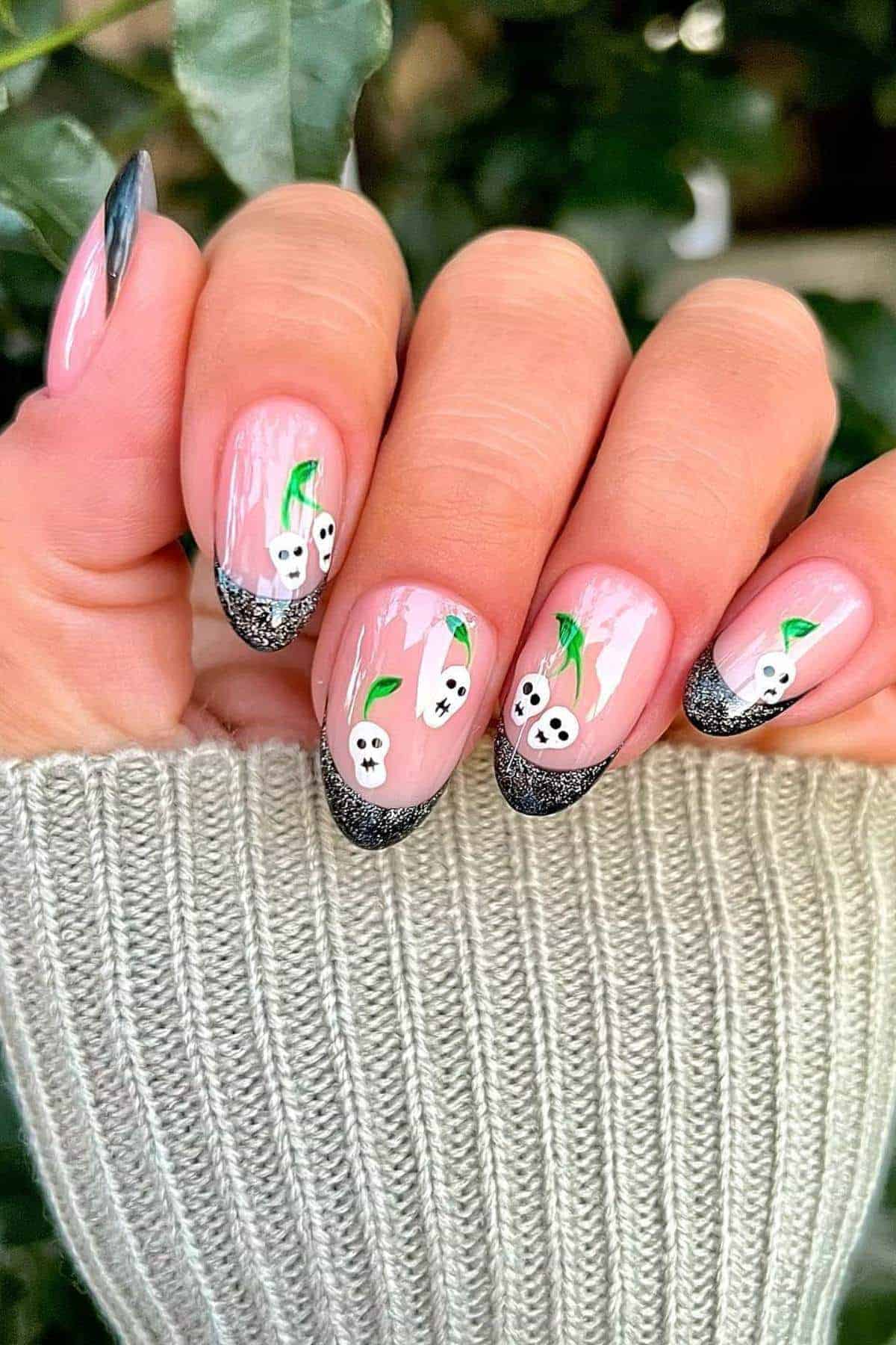 Hand with pink and black glitter-tipped nails featuring small white ghosts and green leaves as nail art designs.